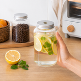 A hand holds a clear glass mason jar filled with lemon slices, mint leaves, and water on a wooden countertop. In the background, two glass storage jars with silver lids sit on a wooden tray next to an orange and a white toaster oven.