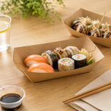 Sushi in a cardboard container on a wooden table with a glass of water and a small bowl.