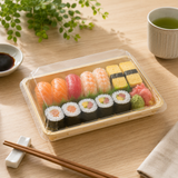 Assorted sushi in a clear container on a wooden table with a cup of green tea and soy sauce.